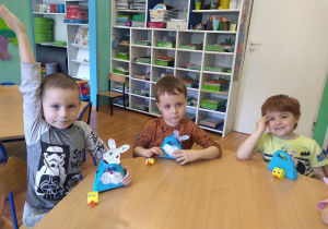 Szymon, Antoś, Kuba prezentują wykonane świąteczne ozdoby. Trzymają zrobione z papieru koszyczki wielkanocne. Na każdym z nich baranek wielkanocny (głowa wycięta z papieru, tułów wyklejony watą) Z koszyczka wystaje zajączek wielkanocny ( głowa wycięta z papieru i pomalowana kredkam a następnie przymocowana do patyczka do szaszłyków ). Obok koszyczka kurczak wykonany z paska żółtego papieru, dziób doklejony z kawałka czerwonego papieru