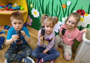 Kuba, Monika i Helenka prezentują prezenty dla Ziemi wykonane z okazji jej święta z materiałów recyklingowych. Rolki od papieru toaletowego pomalowane w kolorze czarnym. Doklejona papierowa głowa kota wycięta i pomalowana na czarno.