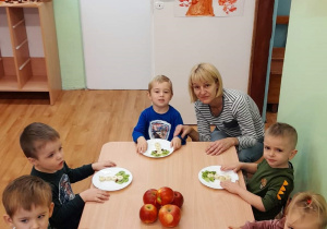 Dzieci siedzą przy stoliku, jedzą przygotowane przez siebie owocowe smakołyki. Przy stoliku z dziećmi jest wychowawczyni.