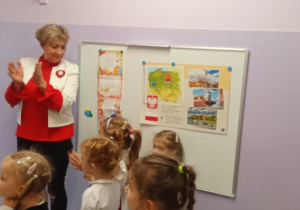 Pani Dyrektor stoi w sali, klaszcze w ręce dziękując dzieciom za udział w akcji Hymn dla Niepodległej. Obok grupka dzieci w strojach galowych