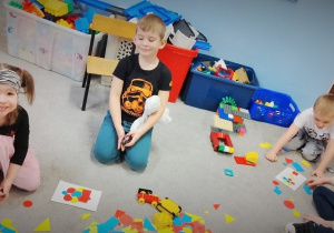 Dzieci siedzą na dywanie. Układają kompozycje z figur geometrycznych według wzorów podanych na kartce.