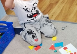 Dzieci siedzą na dywanie. Układają kompozycje z figur geometrycznych według wzorów podanych na kartce.