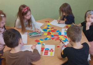 Dzieci siedzą przy stolikach. Przed nimi na kartkach rysunki z figur geometrycznych. Zadaniem przedszkolaków jest odrysowanie ich na swojej kartce zgodnie ze wzorem.