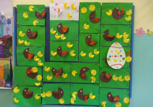 Na dużej tablicy korkowej wywieszone prace dzieci. Kura w kolorze brązowym i żółte kurczęta ( wykonane z kół).