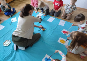 Dzieci siedzą w kole i malują farbami rysunki pisanek.