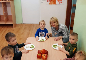 Dzieci siedzą przy stoliku, jedzą przygotowane przez siebie owocowe smakołyki. Przy stoliku z dziećmi jest wychowawczyni.