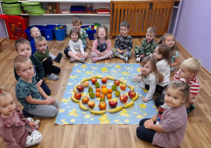Dzieci siedza na podłodze w kole. w środku ułożona przez przedszkolaków kompozycja - grejffrut otoczony kołem z mandarynek. Mandarynki otoczone kołem z gruszek, koło z gruszek otoczone jabłkami, jabłka otoczone kołem z bananów.