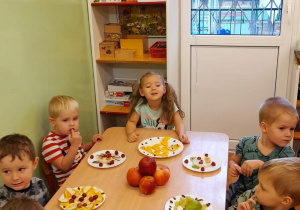 Dzieci siedzą przy stoliku, jedzą przygotowane przez siebie owocowe smakołyki.