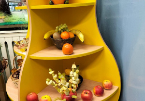 Półka w kształcie gruszki. Na najwyższym poziomie znajduje się doniczka dynia z kompozycją z jesiennych liści i gałązek. Na niższej półce misa wypełniona owocami: bananami, winogronem, kiwi, pomarańczami. Na trzeciej półce od góry szaszłyki owocowe wbite w jabłko. Obok jabłka z szaszłykami, ułożone jabłka - jedno obok drugiego. Na najniższej półce książka: Co nam daje las