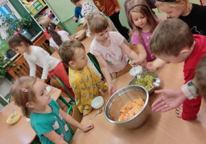 Dzieci stoją przy stolikach na których przygotowane są składniki do wykonania sałatki owocowej